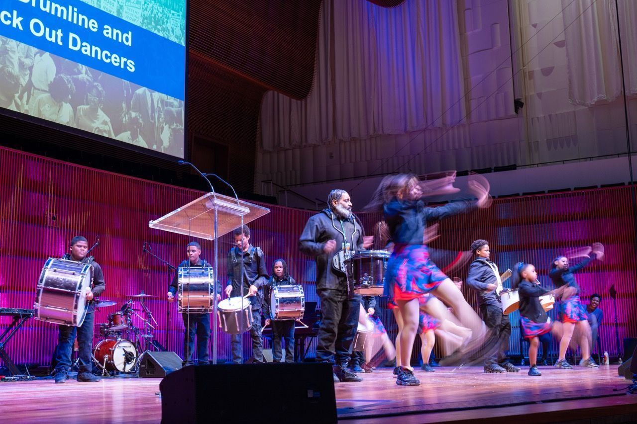 A group of children and an adult performing on stage with drums and dancers. The dancers in colorful skirts are in motion towards the right, creating a blurred effect. The drummers stand in a line behind a clear podium with microphones, set against a purple-lit auditorium backdrop. A large screen above shows partially visible white text on a blue background.